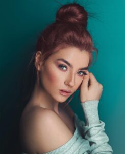 A fashionable adult woman posing with elegant makeup against a teal background.
